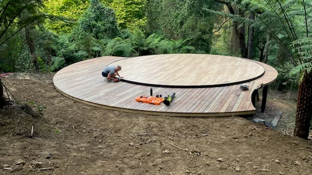 Person working on a large circular wooden yurt platform in a forest setting