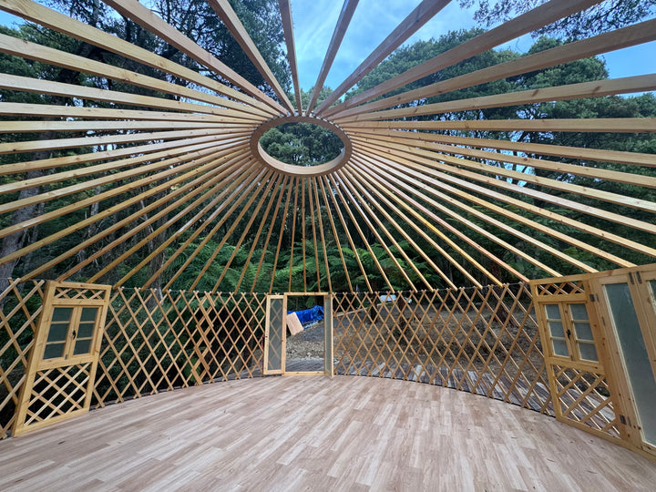 Wooden yurt structure with lattice walls and floor, surrounded by trees.