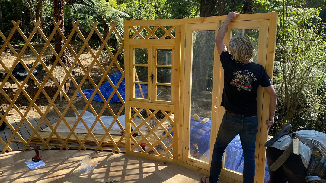 Person assembling a wooden yurt structure outdoors with tools and materials around.
