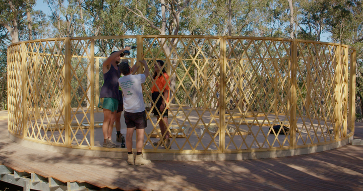 Three people working on wooden yurt lattice walls in a natural setting.