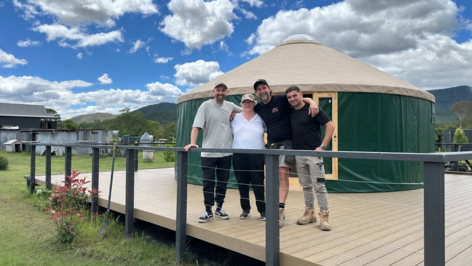 A small group standing on the deck in front of a finished Beyond Yurt, with green walls, a beige roof and mountain views behind.