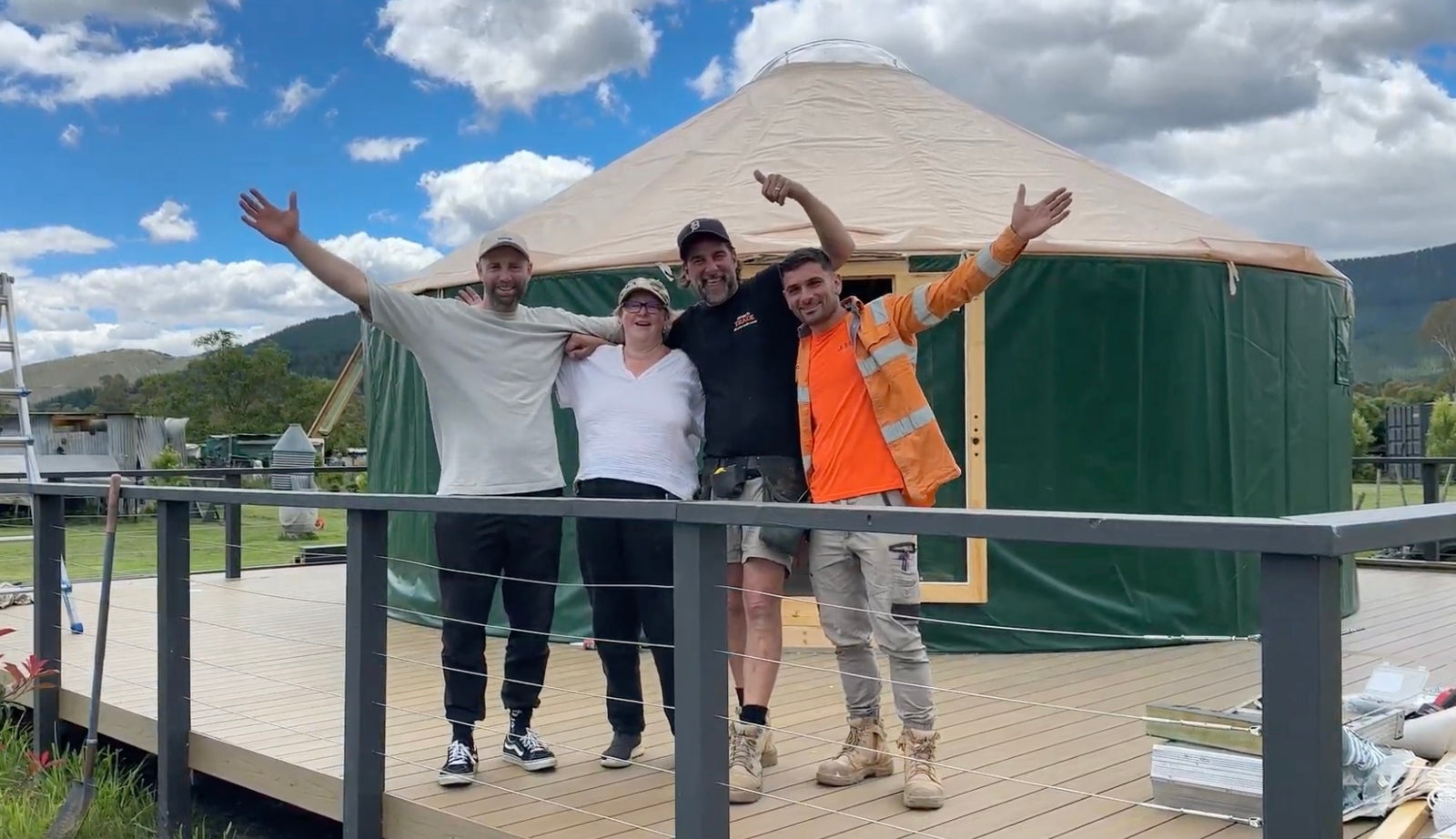 A small group standing on the deck in front of a finished Beyond Yurt, with green walls, a beige roof and mountain views behind.